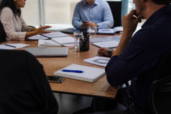 Sala de reuniões com mesa, cadeiras e documentos, pessoas de costas ou perfil em discussão, sem rostos identificáveis