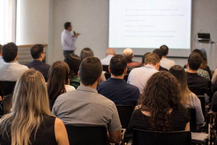 Sala de palestra com plateia de costas observando palestrante diante de projetor, sem rostos identificáveis