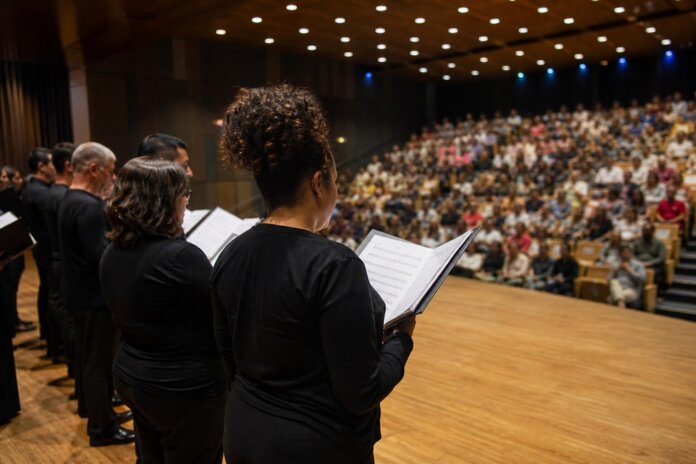 Coral em formação no palco, cantando, com público desfocado ao fundo em plateia, sem rostos identificáveis