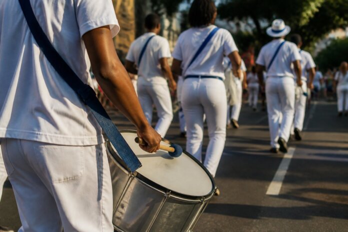 Participantes em desfile caminhando pela avenida, pessoas de costas e ao fundo desfocado, sem rostos identificáveis