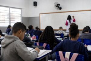 Sala de aula com crianças sentadas às carteiras, professor ao fundo, alto-falante discreto na parede, sem rostos identif